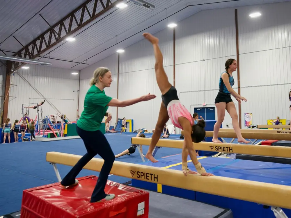 A coach in green spotting a gymnast on the balance beam