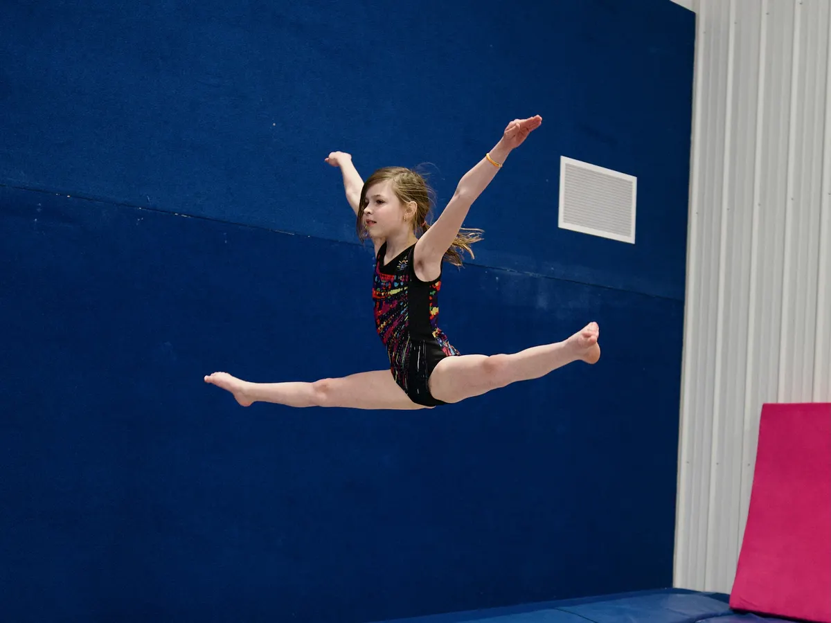 A team gymnast in a full split leap against the blue wall of the tumbling area